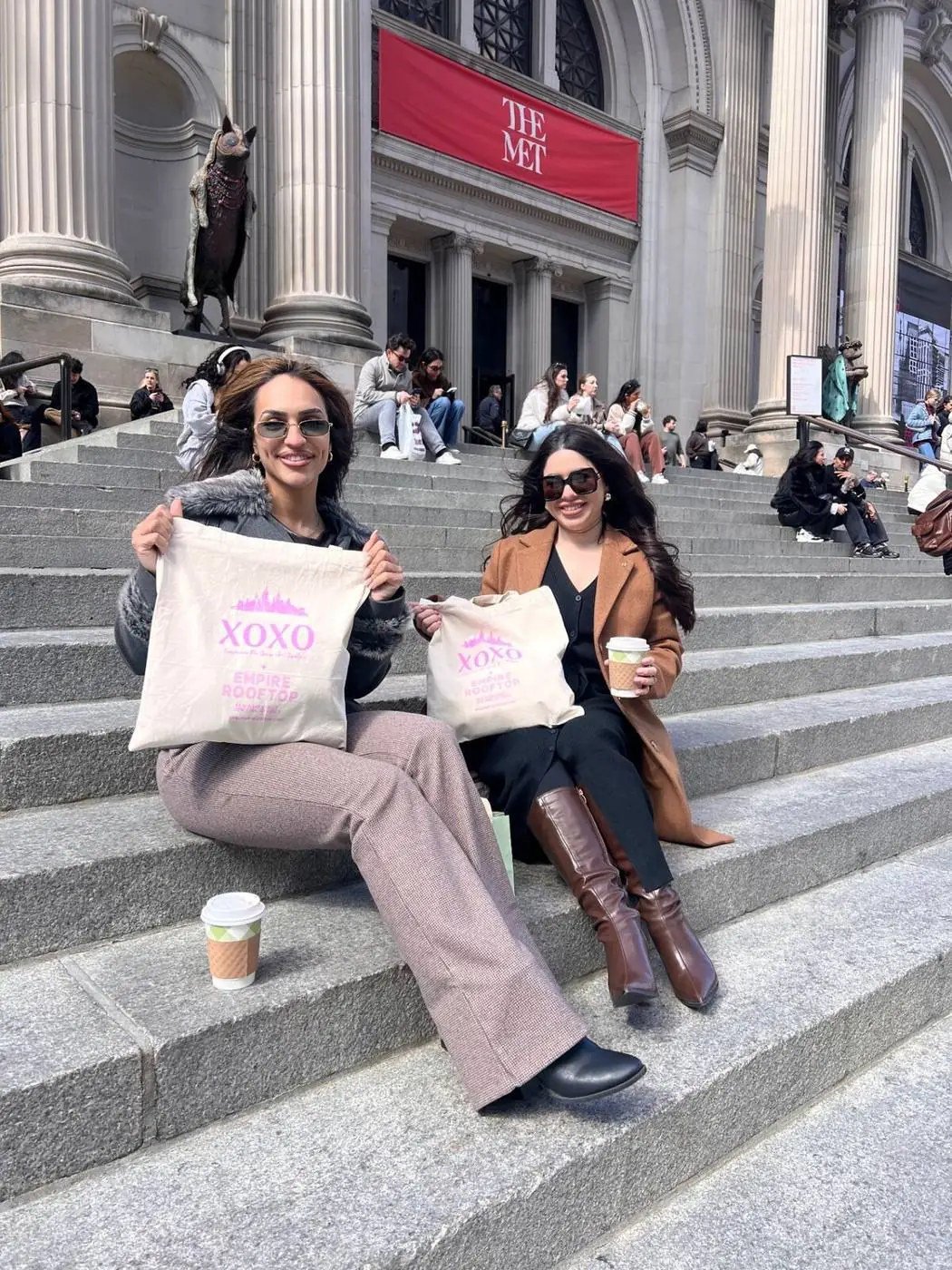Two guests with XOXO tote bags and coffee on the Met Steps